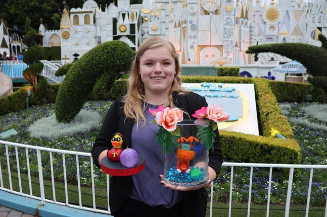 A cast member poses in front of "it's a small world" with two of her rubber duck designs