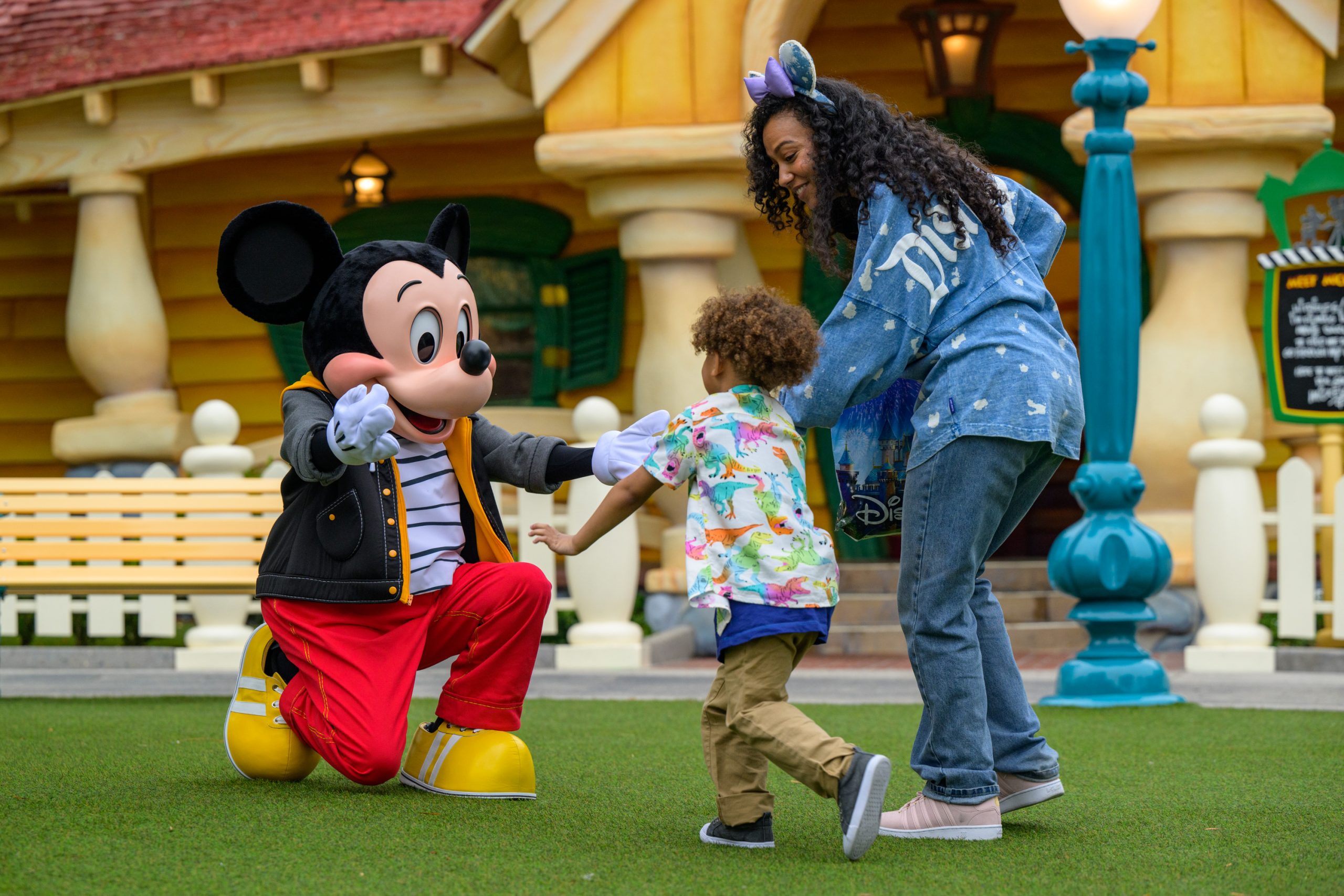 A woman and small child excitedly greet Mickey Mouse in front of his house, with Mickey's arms outstretched to say hi. 