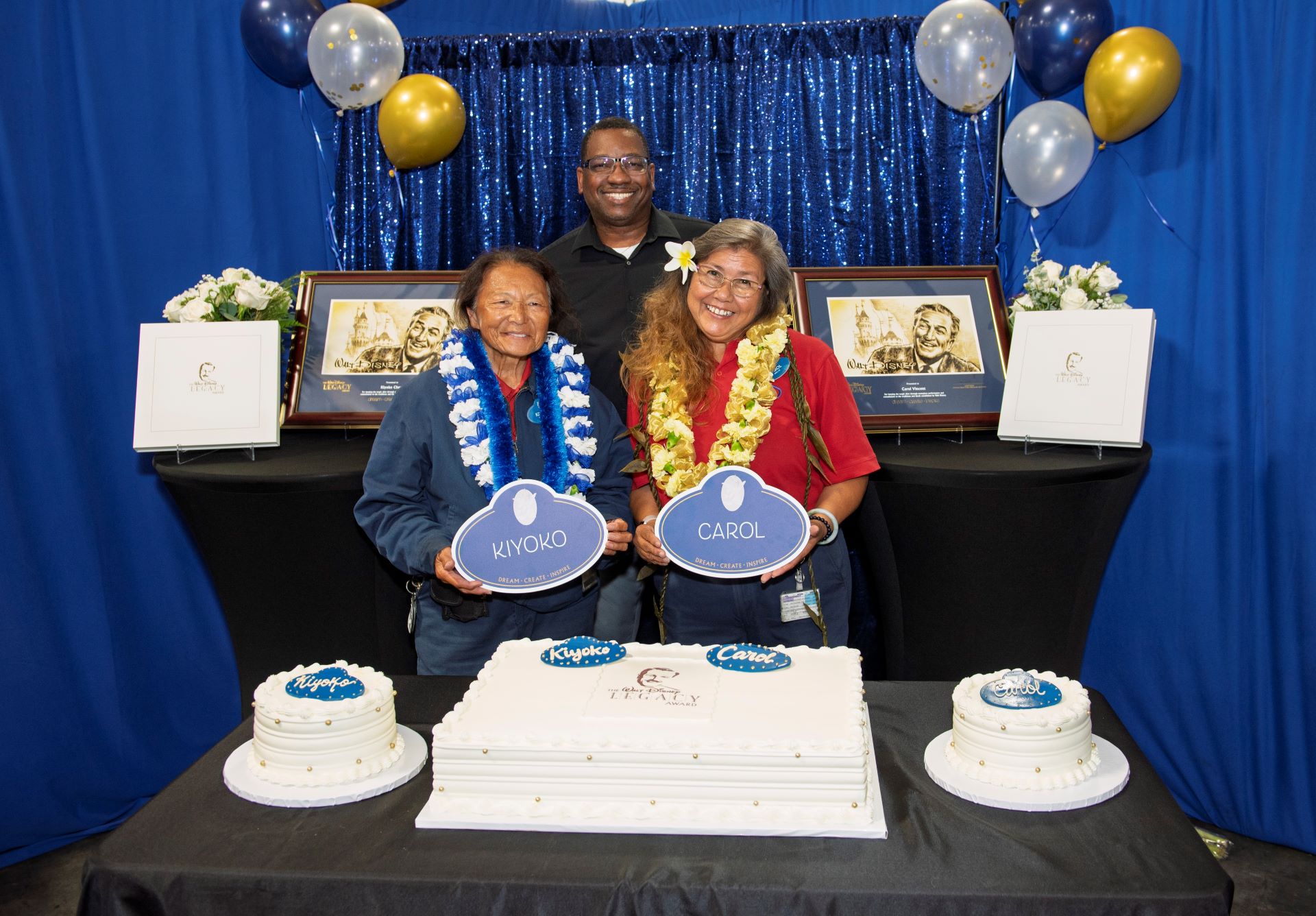 Two women hold award signs, smiling with cakes and decorations behind them.