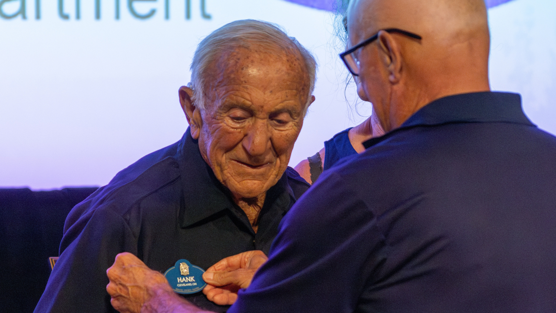 Elderly man receiving a name tag during an award ceremony.
