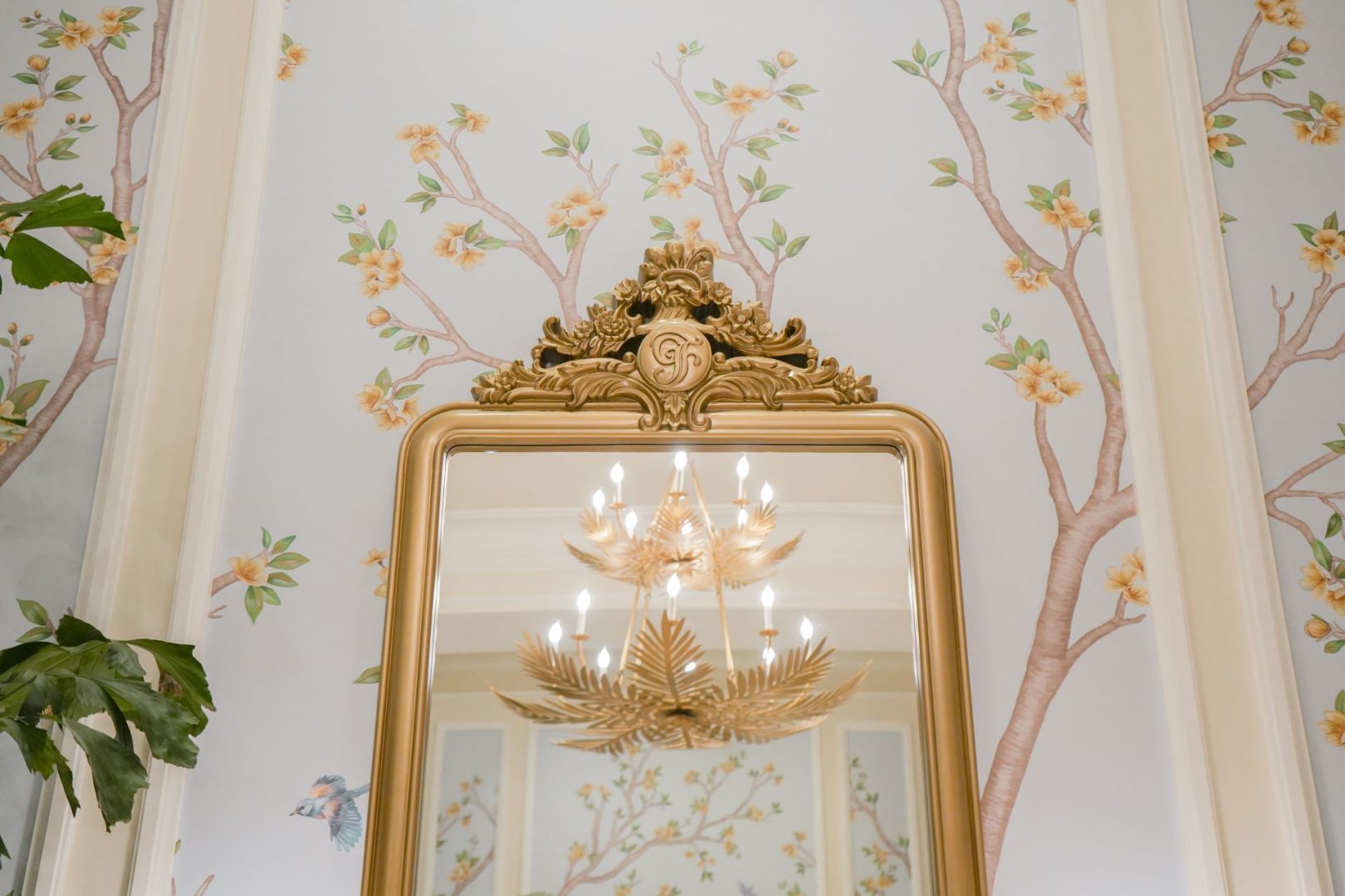 Golden mirror reflecting a chandelier and floral wallpaper in a lobby.