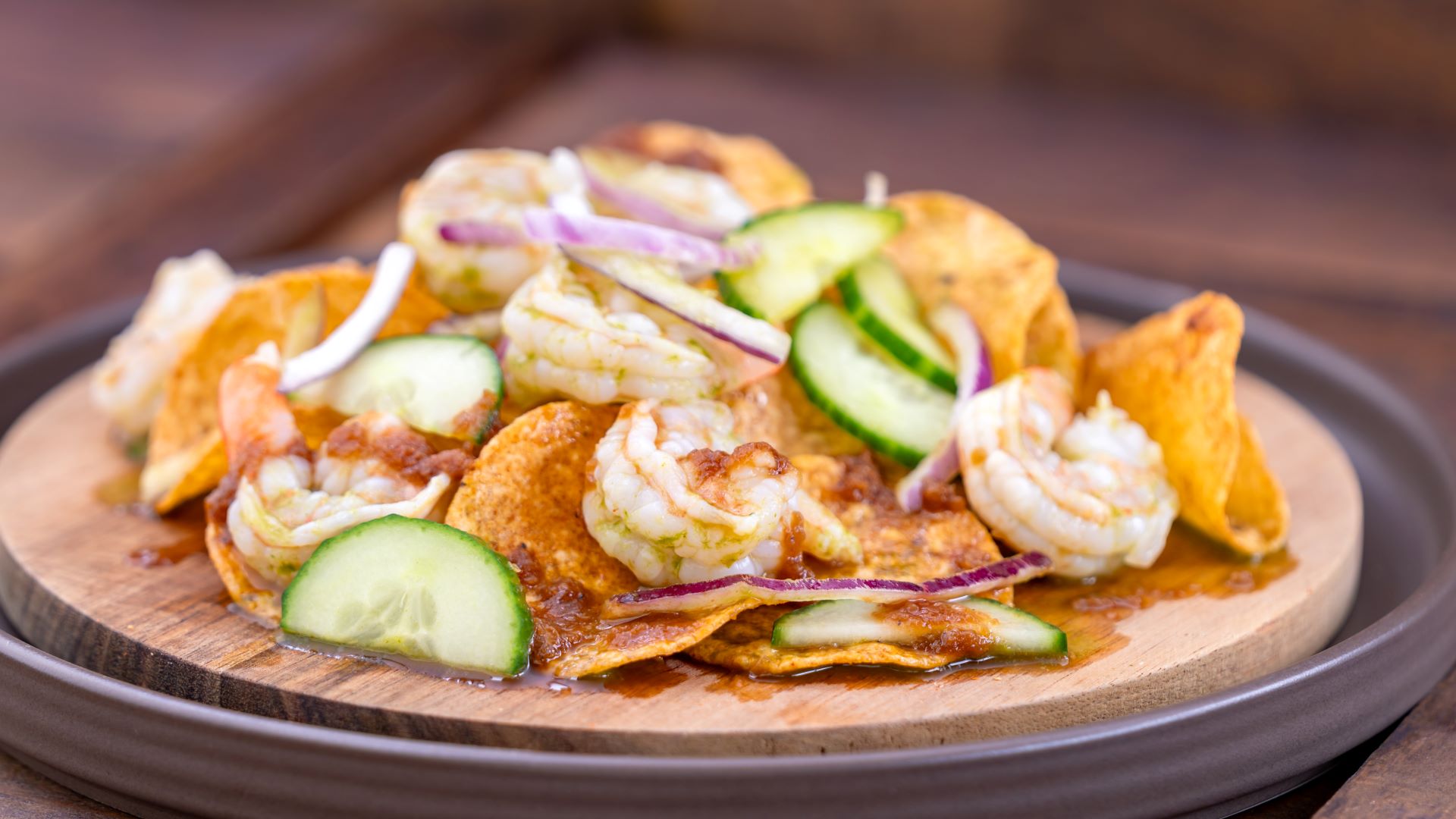 Loaded nachos topped with shrimp, cucumber, and red onion on a wooden plate.