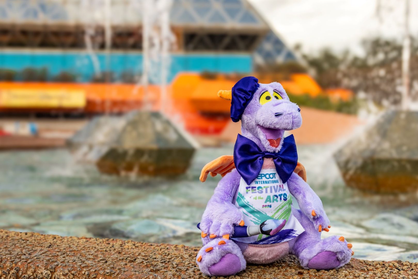 Purple plush dragon wearing a bow tie, in front of a fountain.