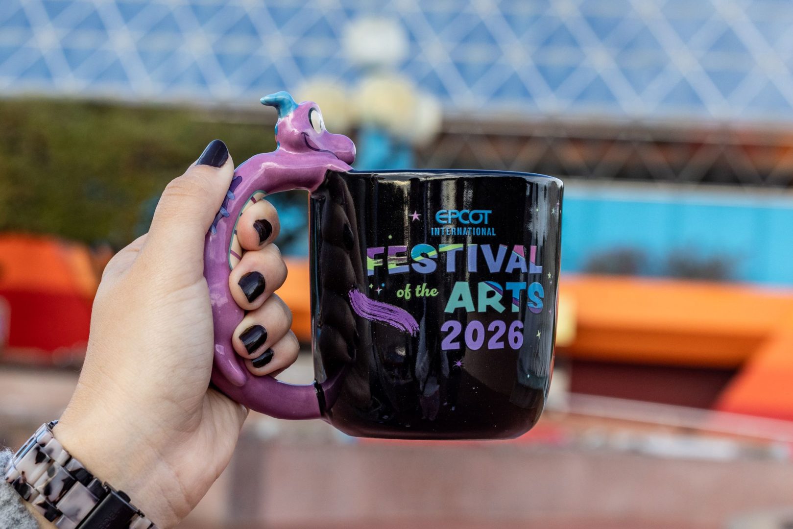 Hand holding a black mug with colorful "Festival of the Arts 2026" design.