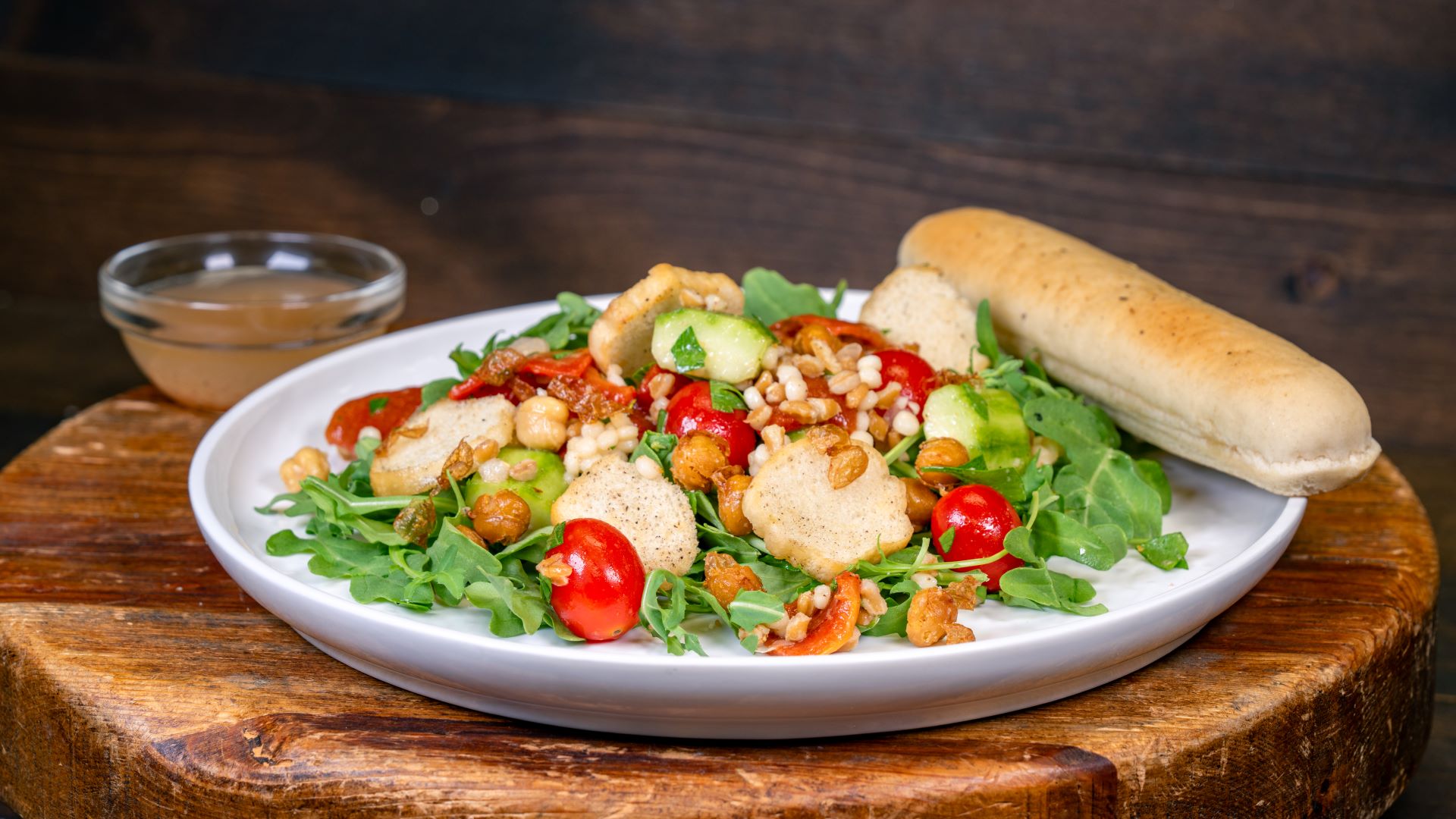 Garden grain salad with tomatoes, cucumbers, and a breadstick on a wooden table.