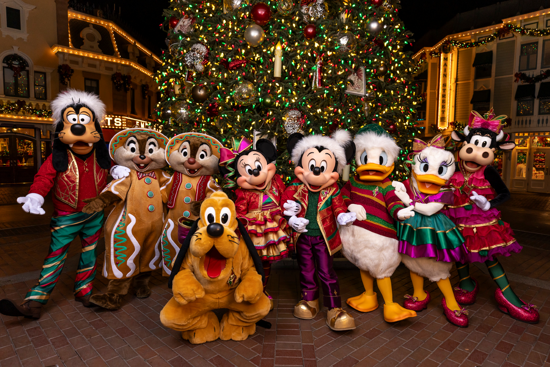 Mickey and friends posing in festive outfits by a Christmas tree.