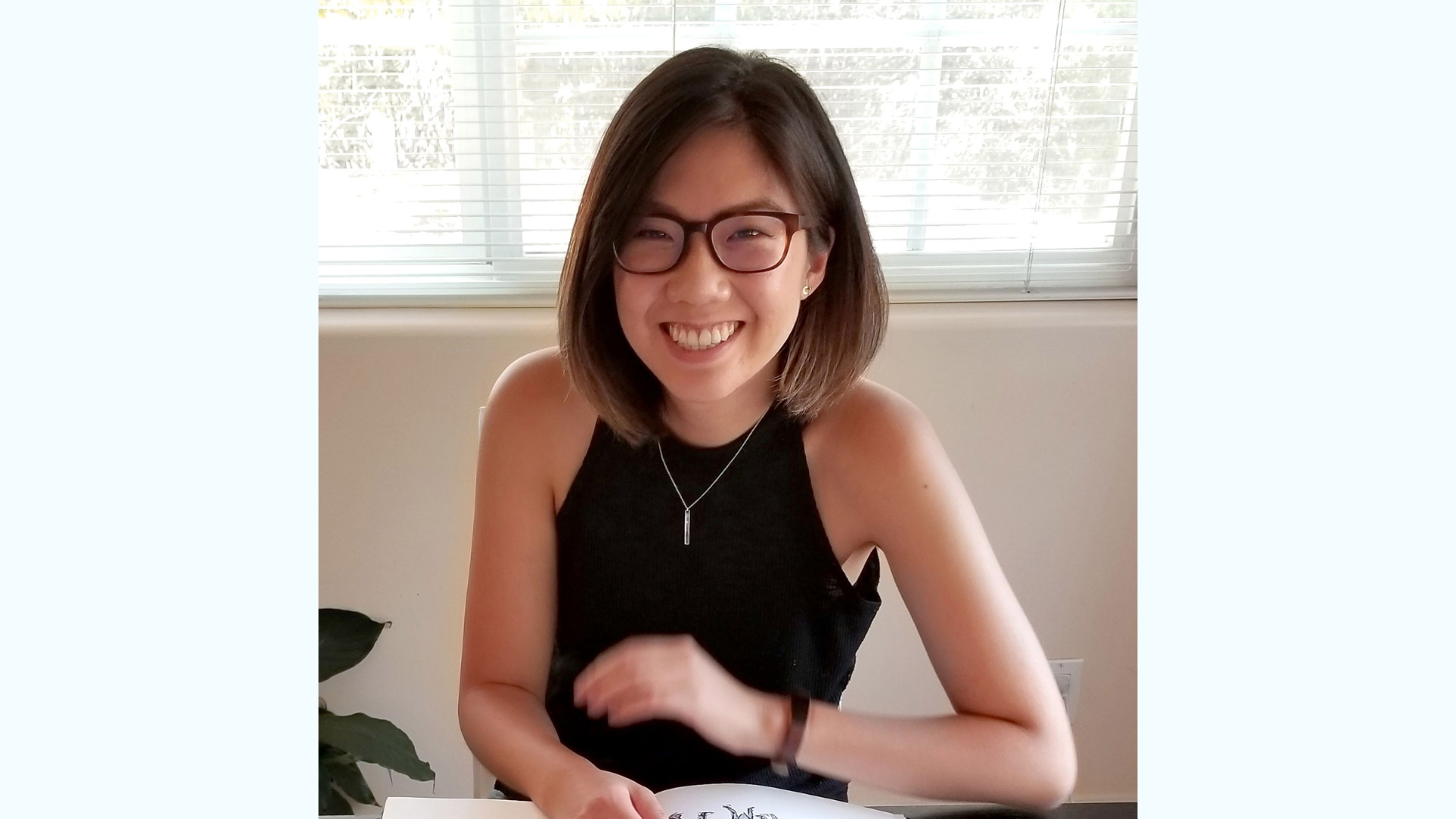 Smiling artist sitting at a table with artwork, indoors with natural light.