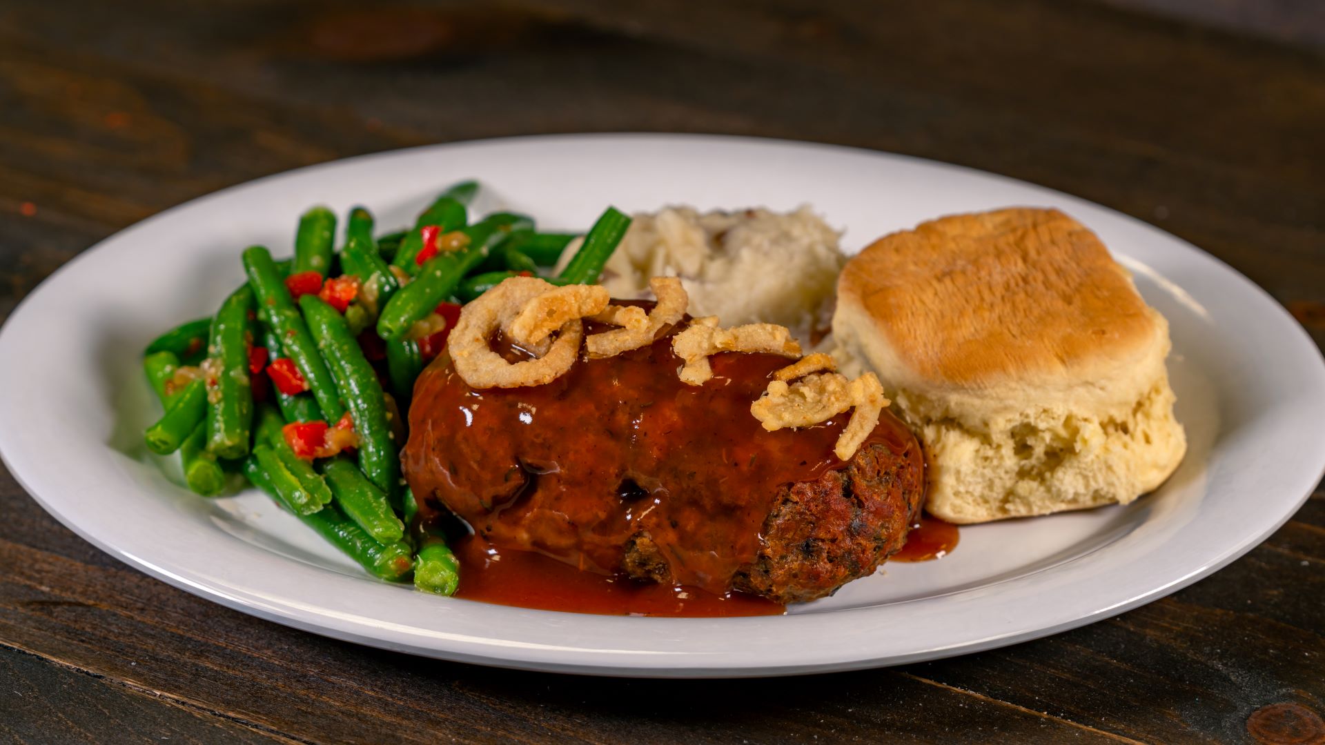 Meatloaf with gravy, green beans, mashed potatoes, and a biscuit on a plate.