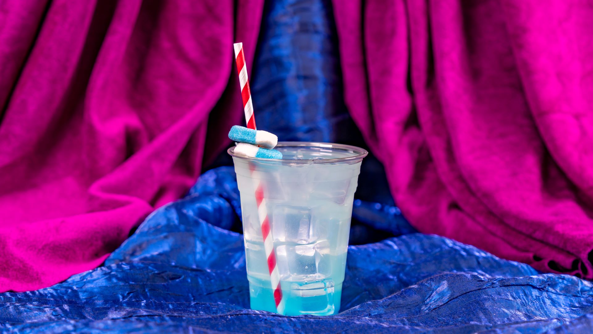 Transparent cup of blue drink with ice and striped straw, draped fabric background.