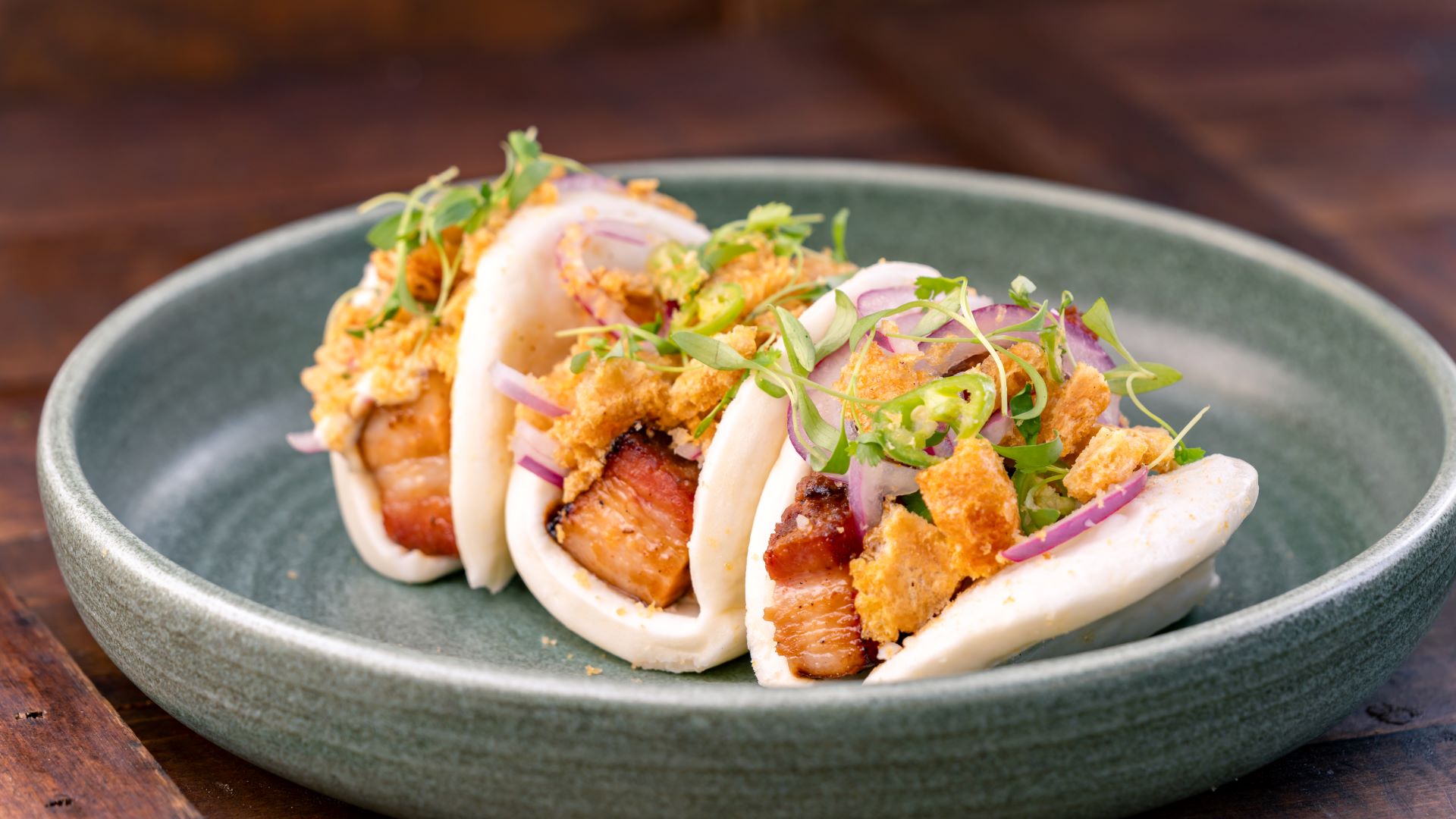 Three bao buns filled with pork belly, garnished with herbs and crispy toppings.