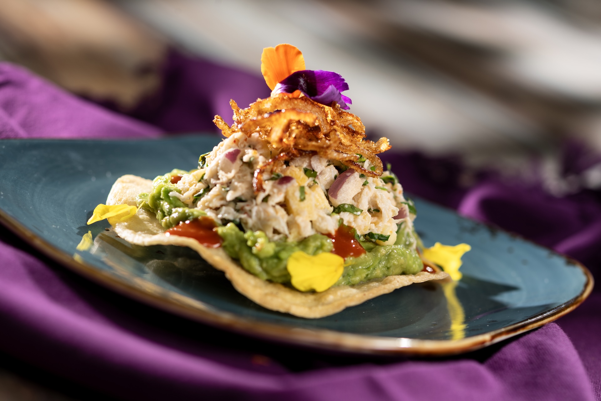 Tostada de Cangrejo: Crispy corn tortilla topped with guacamole, lump crab, mango, crispy onions, and hot sauce (New)