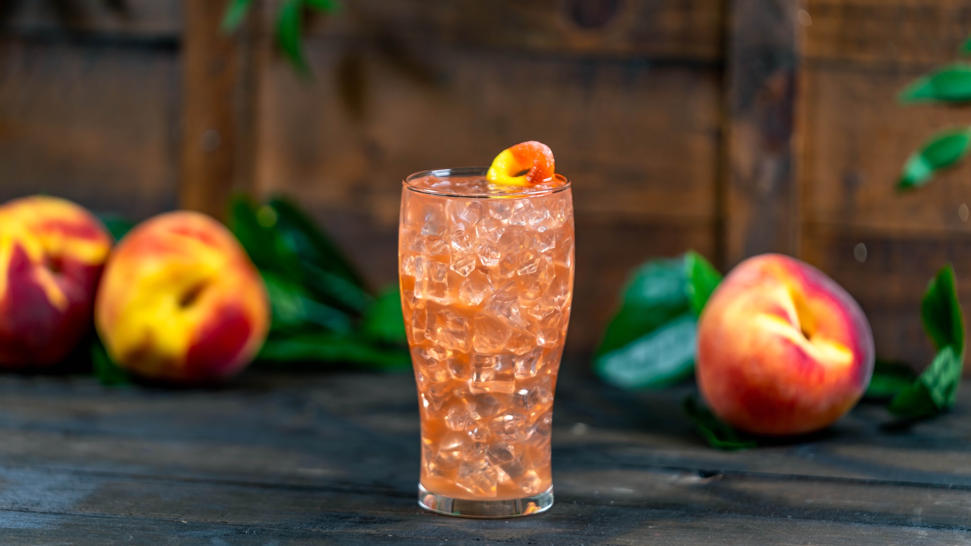 A glass of peach lemonade with ice, surrounded by fresh peaches.
