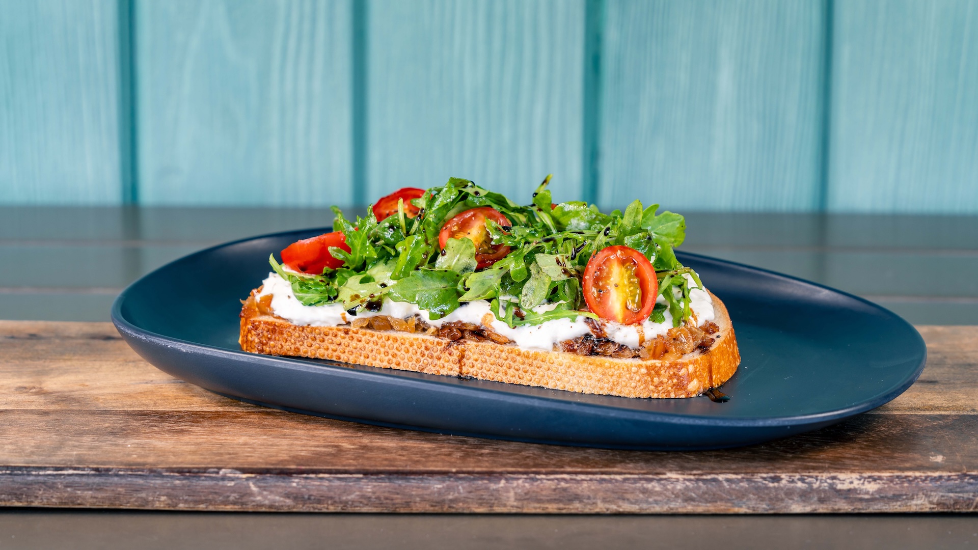 Burrata toast topped with arugula and cherry tomatoes on a dark plate.