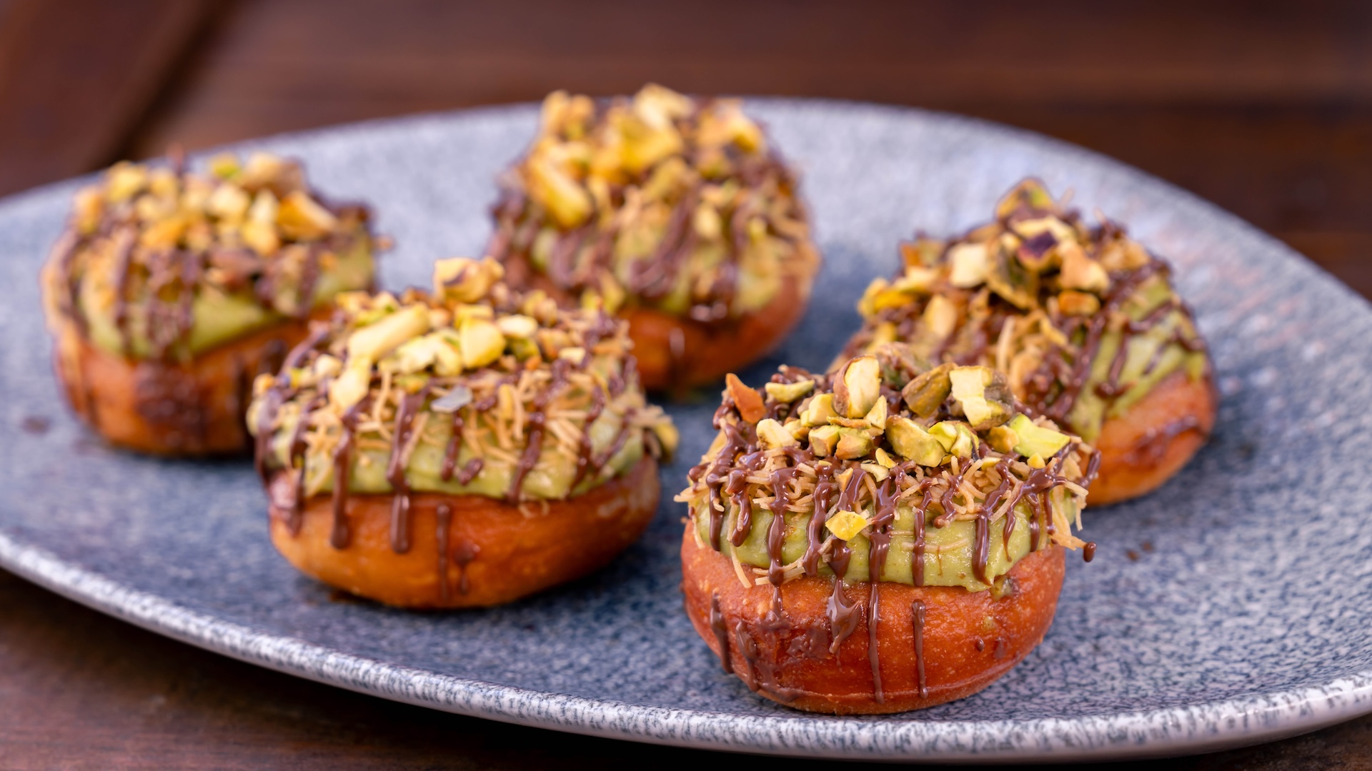 Five chocolate donuts topped with green frosting and crushed pistachios.