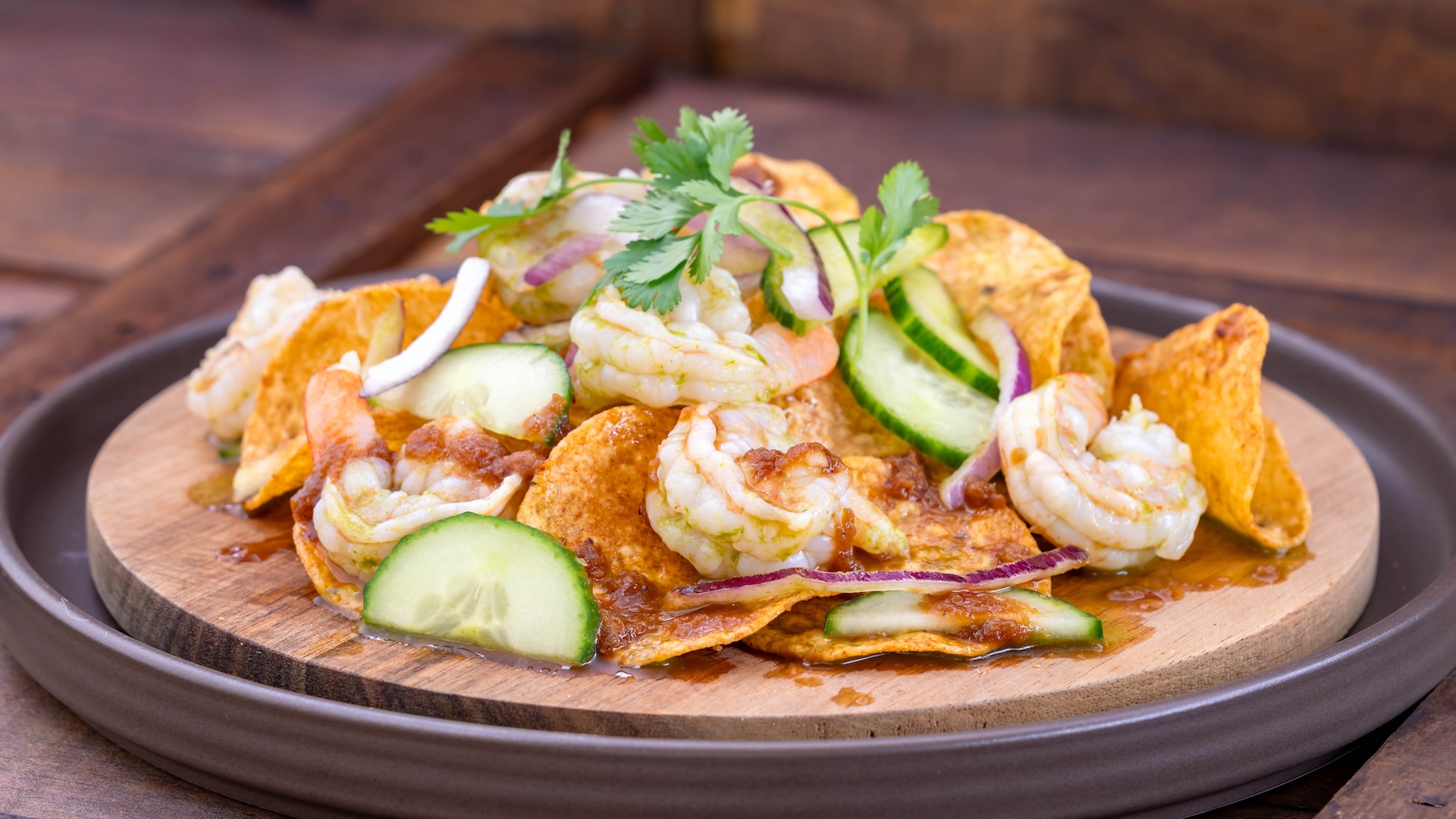 Loaded nachos topped with shrimp, cucumbers, and cilantro on a wooden plate.