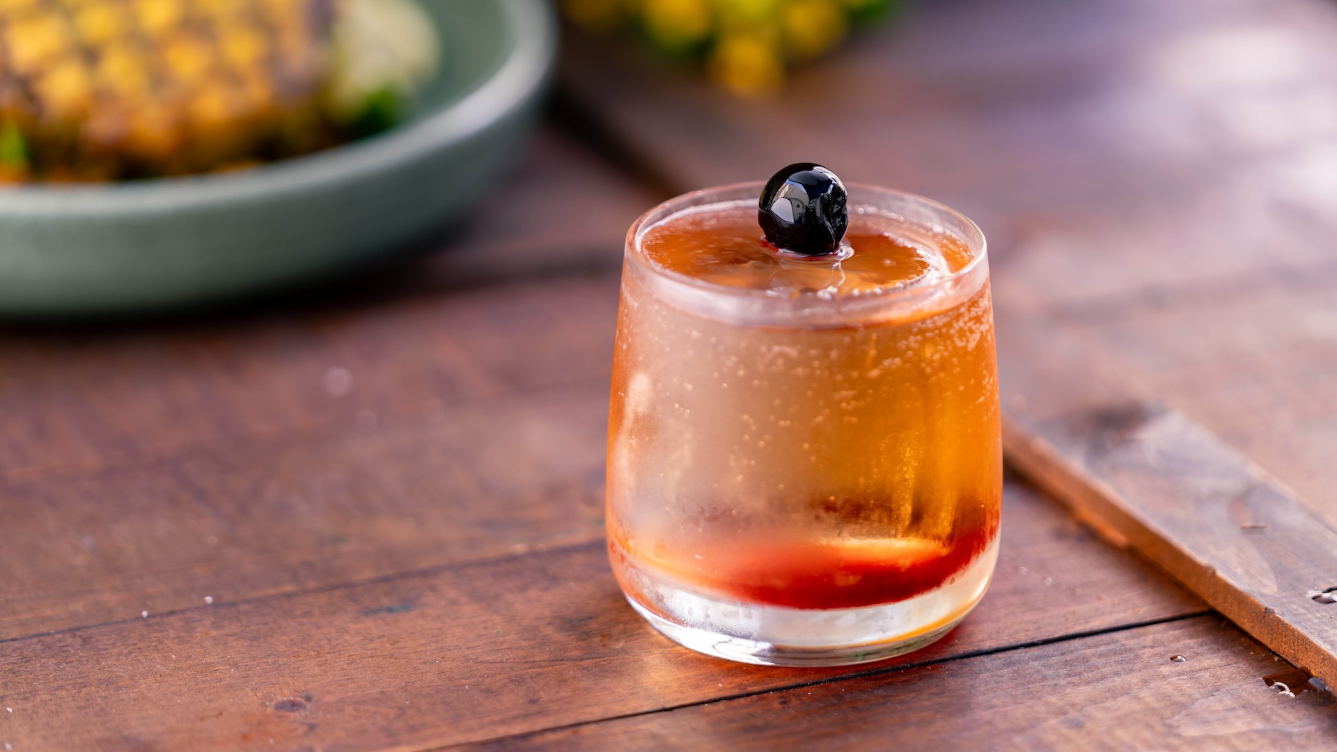Glass of amber-colored cocktail garnished with a cherry on a wooden table.
