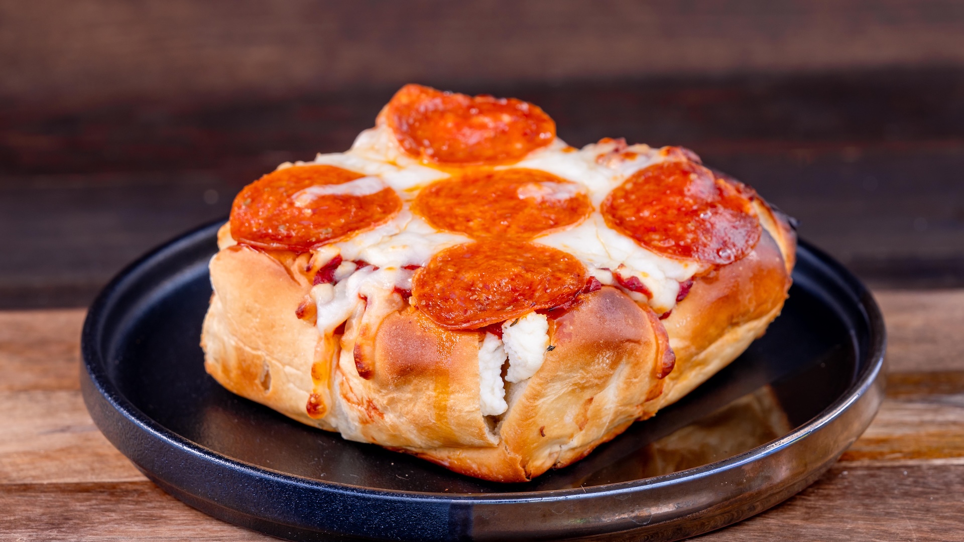 Pepperoni pizza bread on a black plate, set against a wooden background.