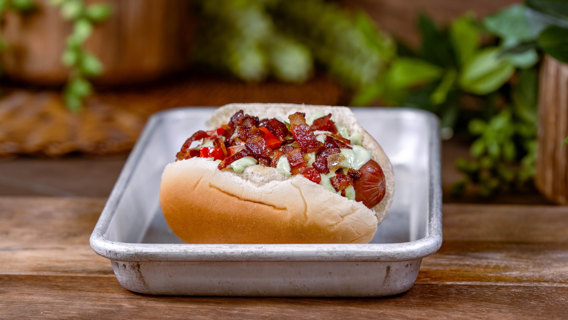 Hot dog topped with bacon, red peppers, and green sauce in a tray.