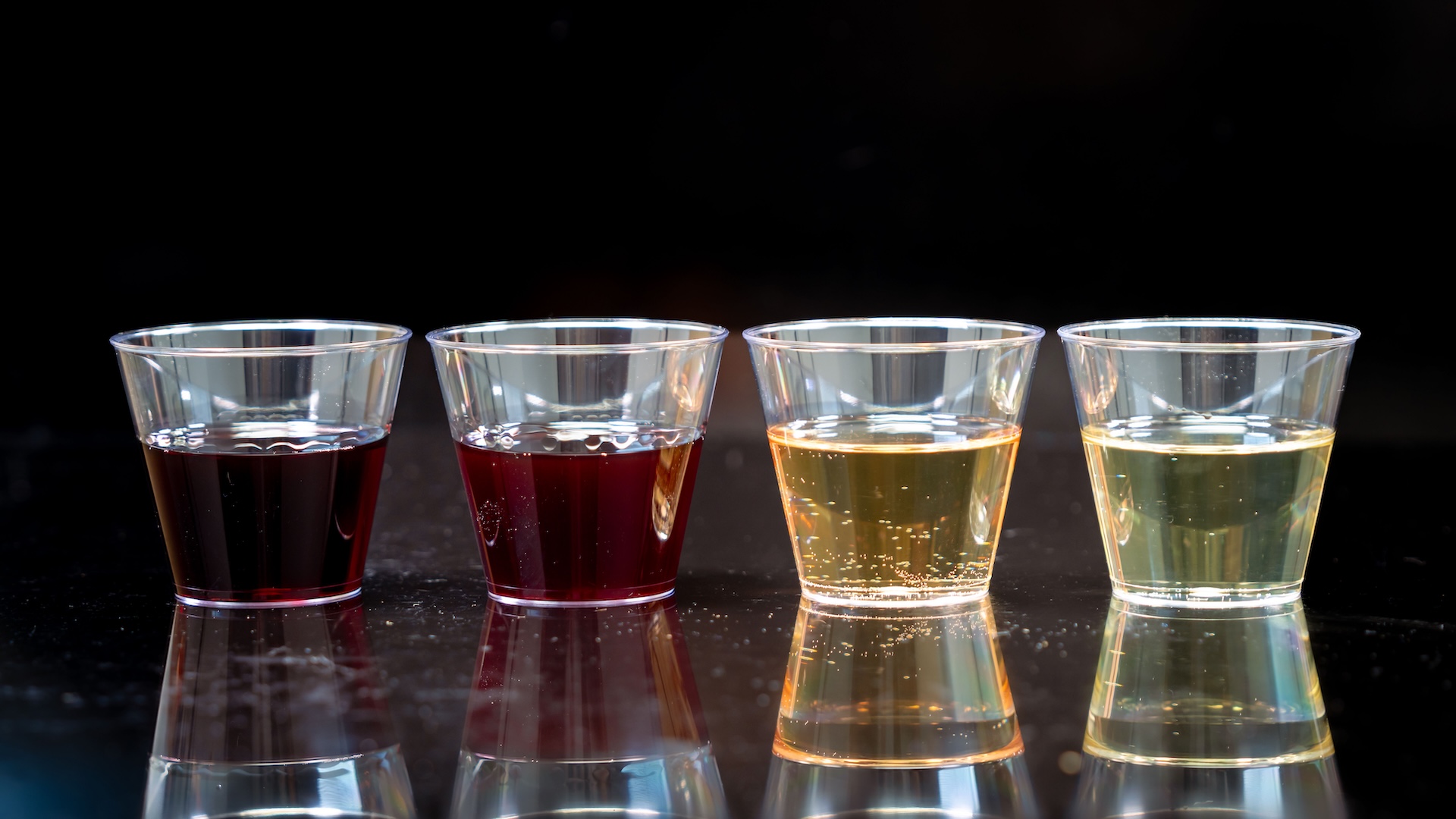 Four plastic cups filled with different colored wines on a reflective surface.