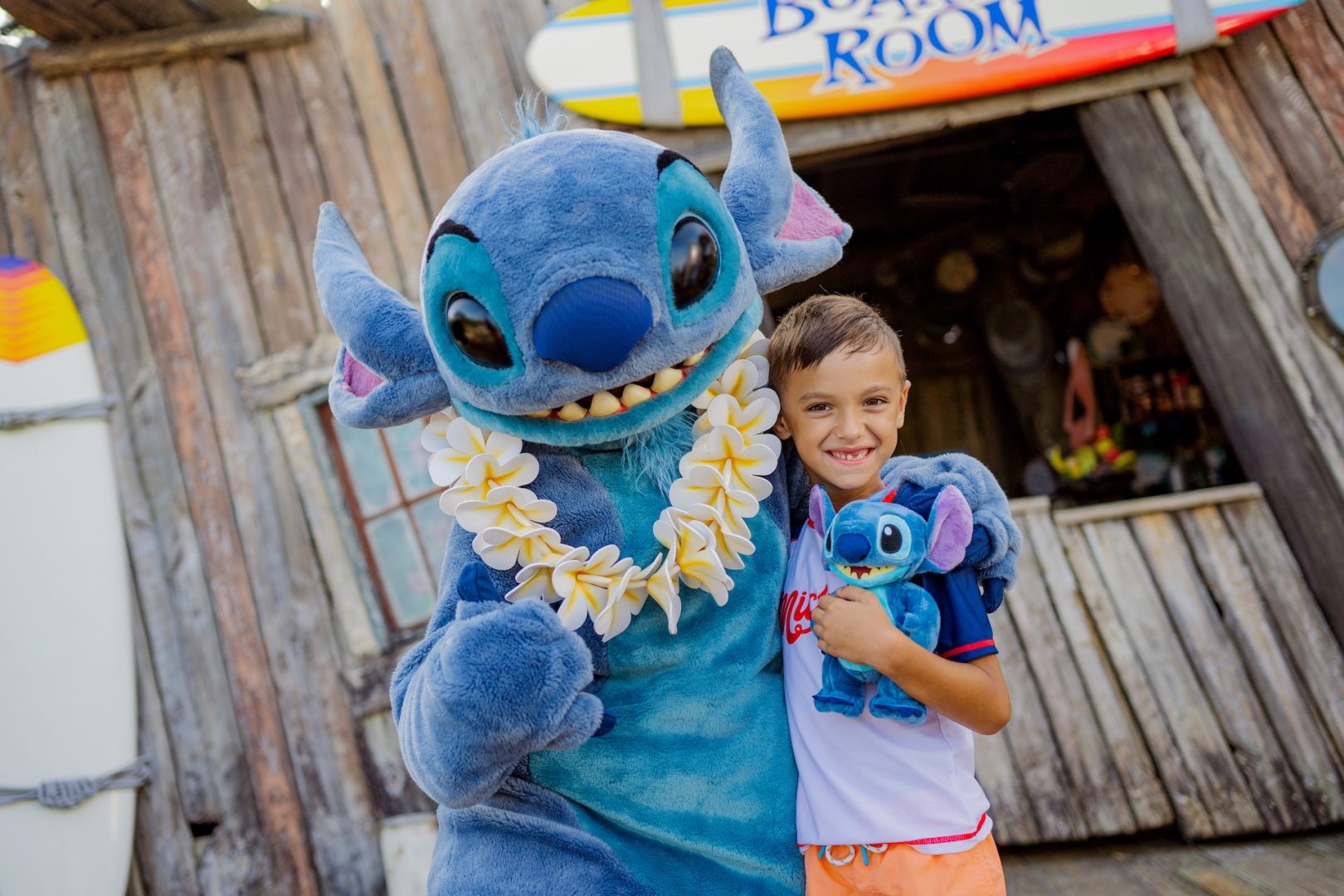 Child poses with Stitch character and a plush toy, tropical setting.