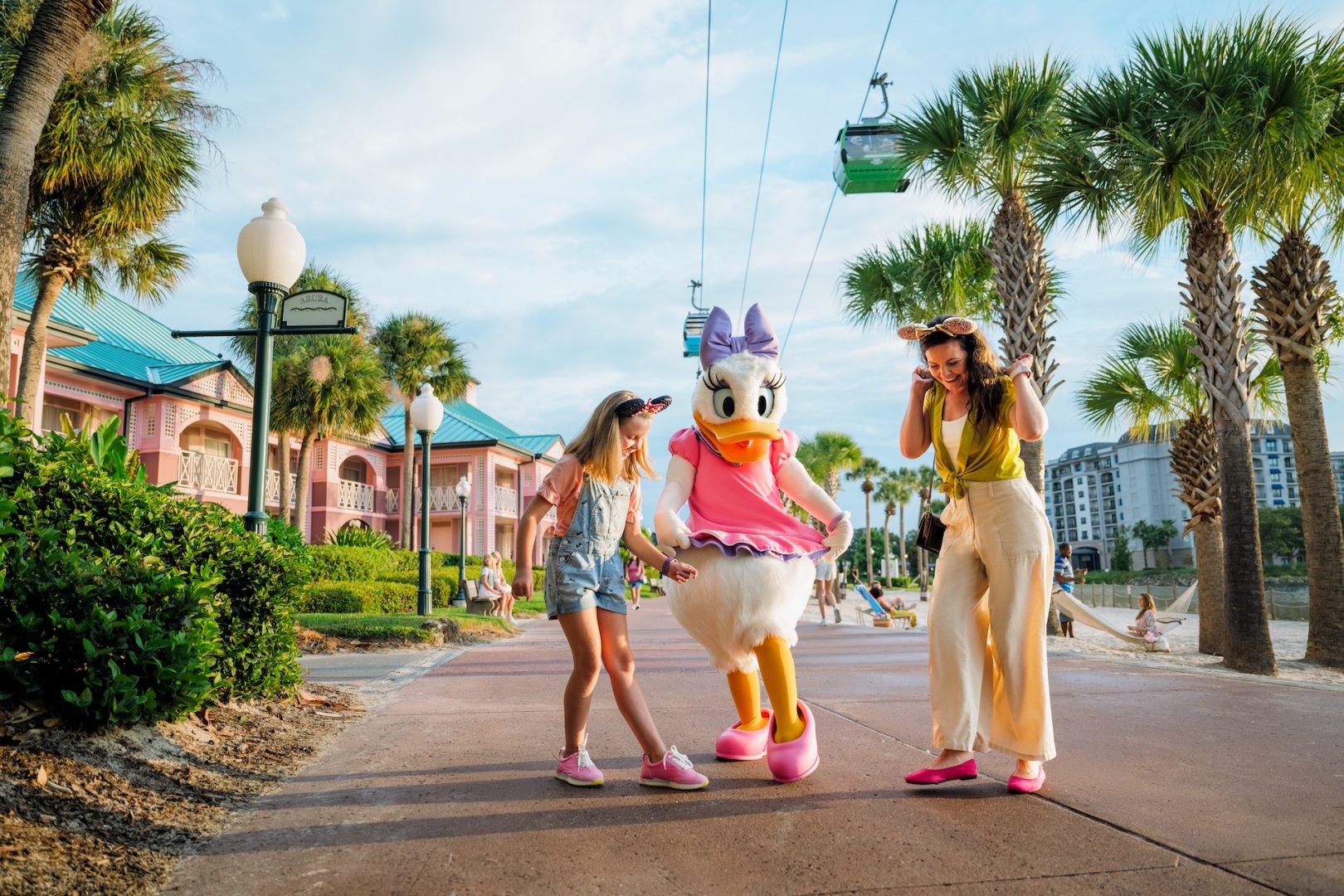 Two girls interact with Daisy Duck near palm trees and colorful buildings.