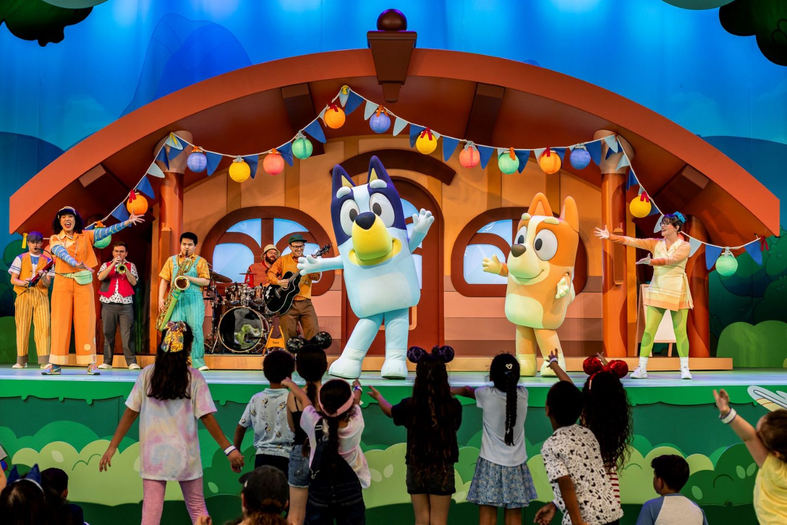 Bluey and Bingo perform on stage with musicians, entertaining children in front.