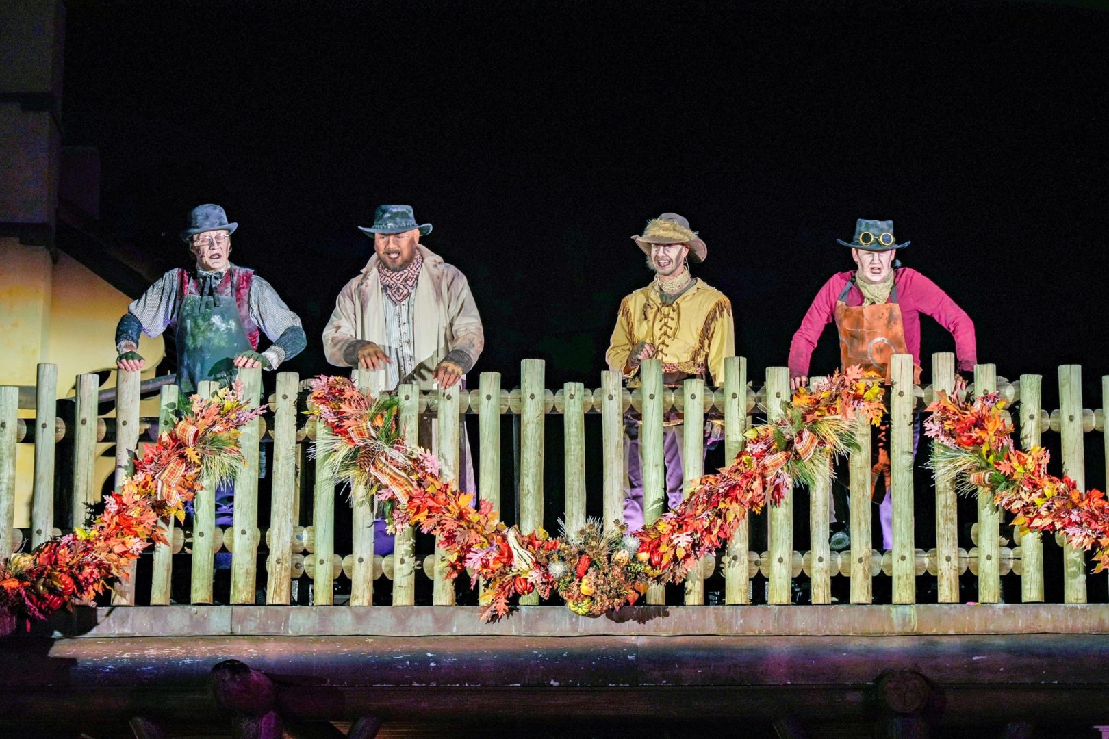 Four costumed performers on a wooden balcony decorated with autumn foliage.