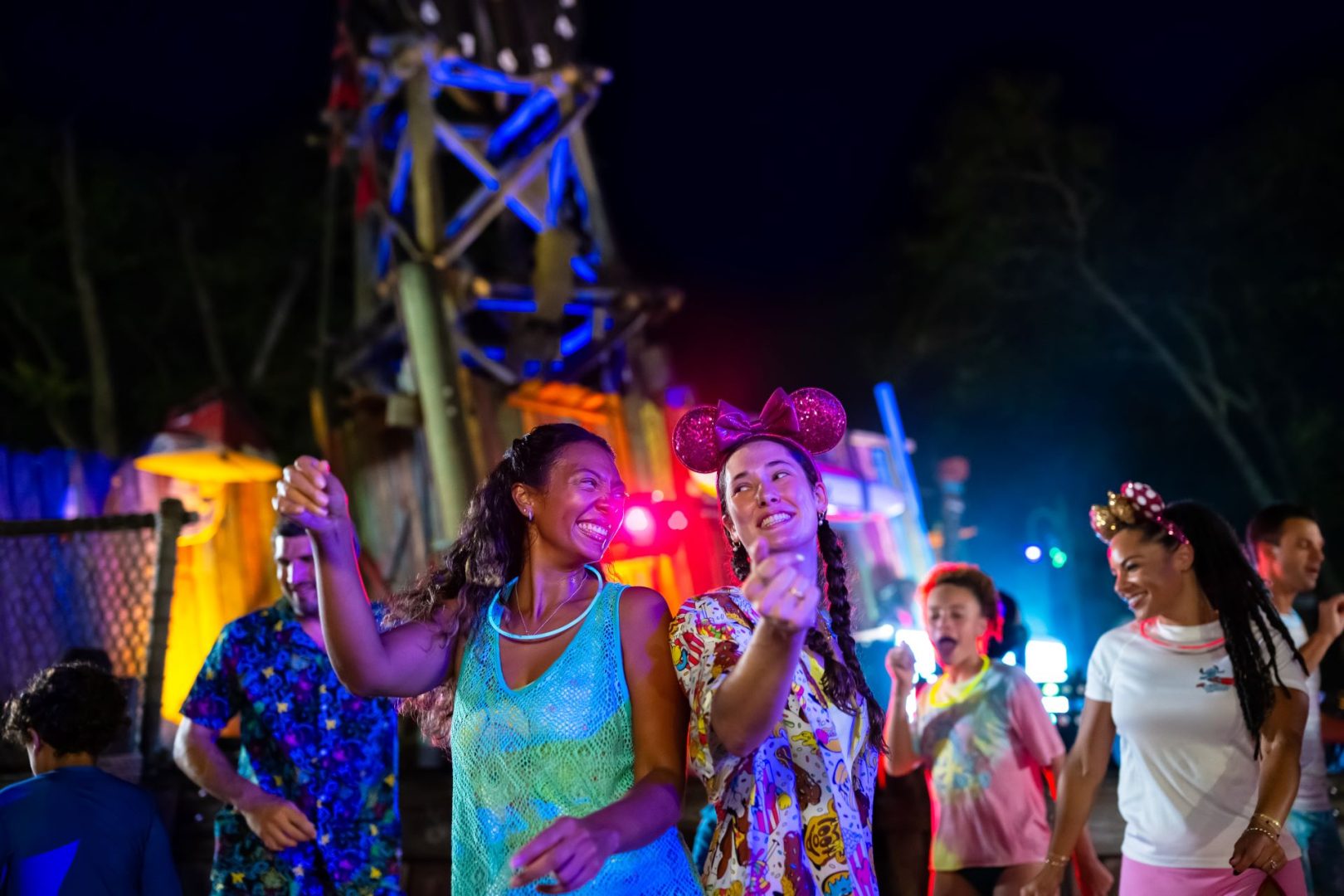 Two women dance joyfully at a colorful nighttime event with friends.