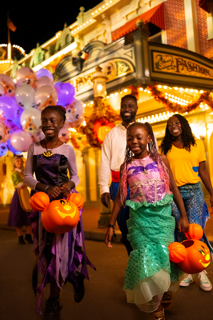 Children in costumes holding pumpkin treat bags walk in a festive setting.