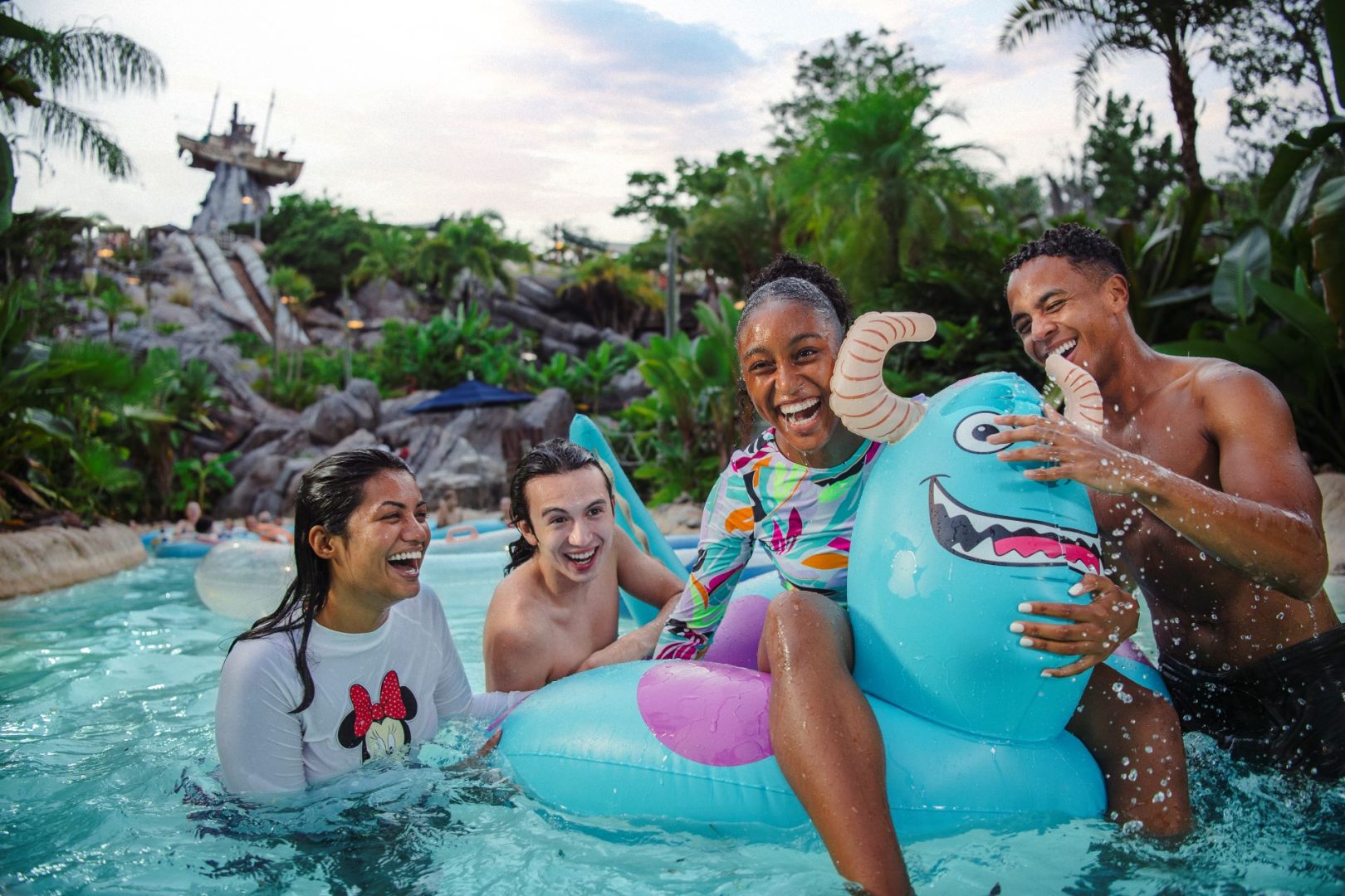 Four friends laughing in a water park pool with a blue monster float.