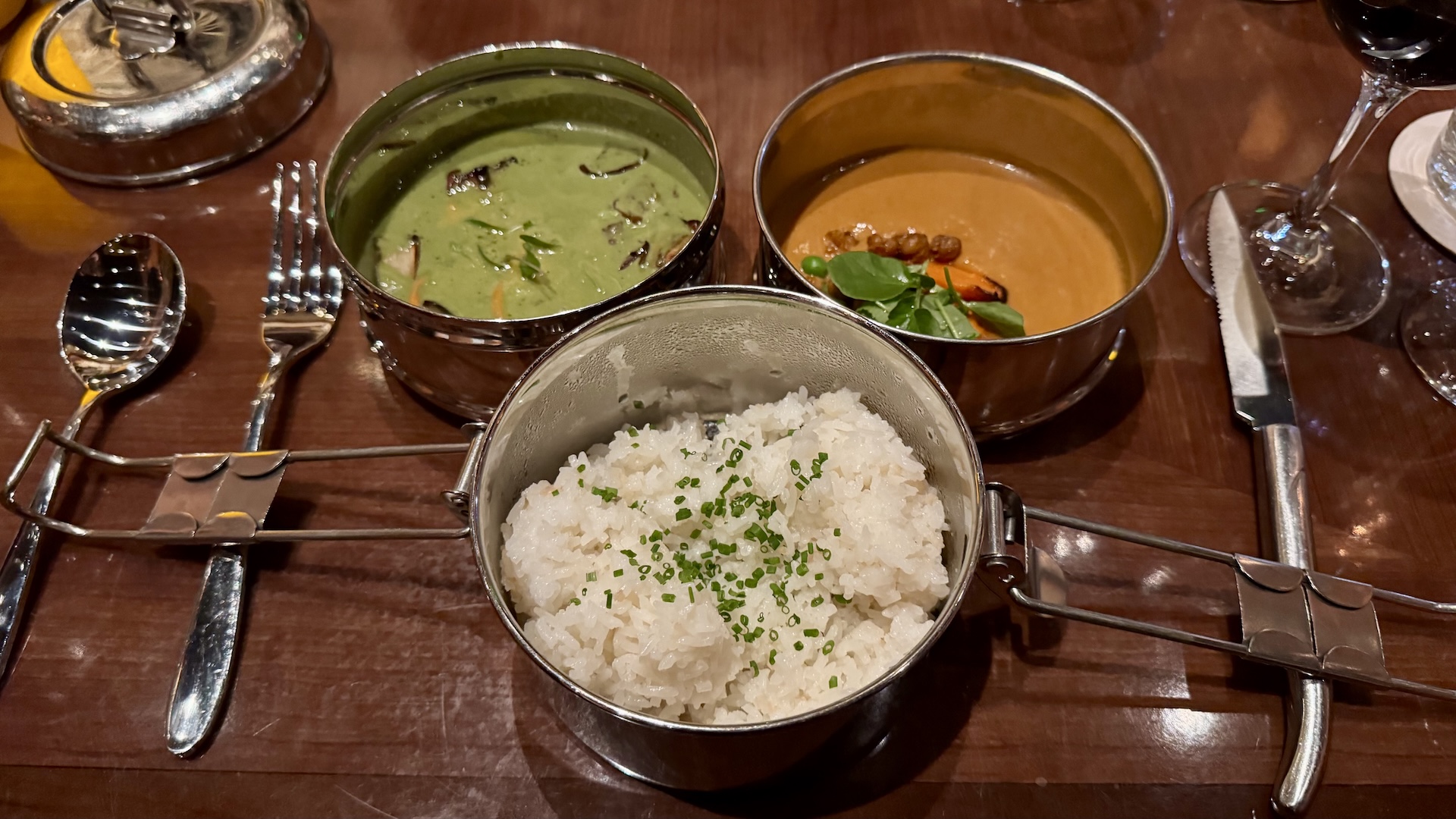 Two curries and rice served in metal containers on a wooden table.