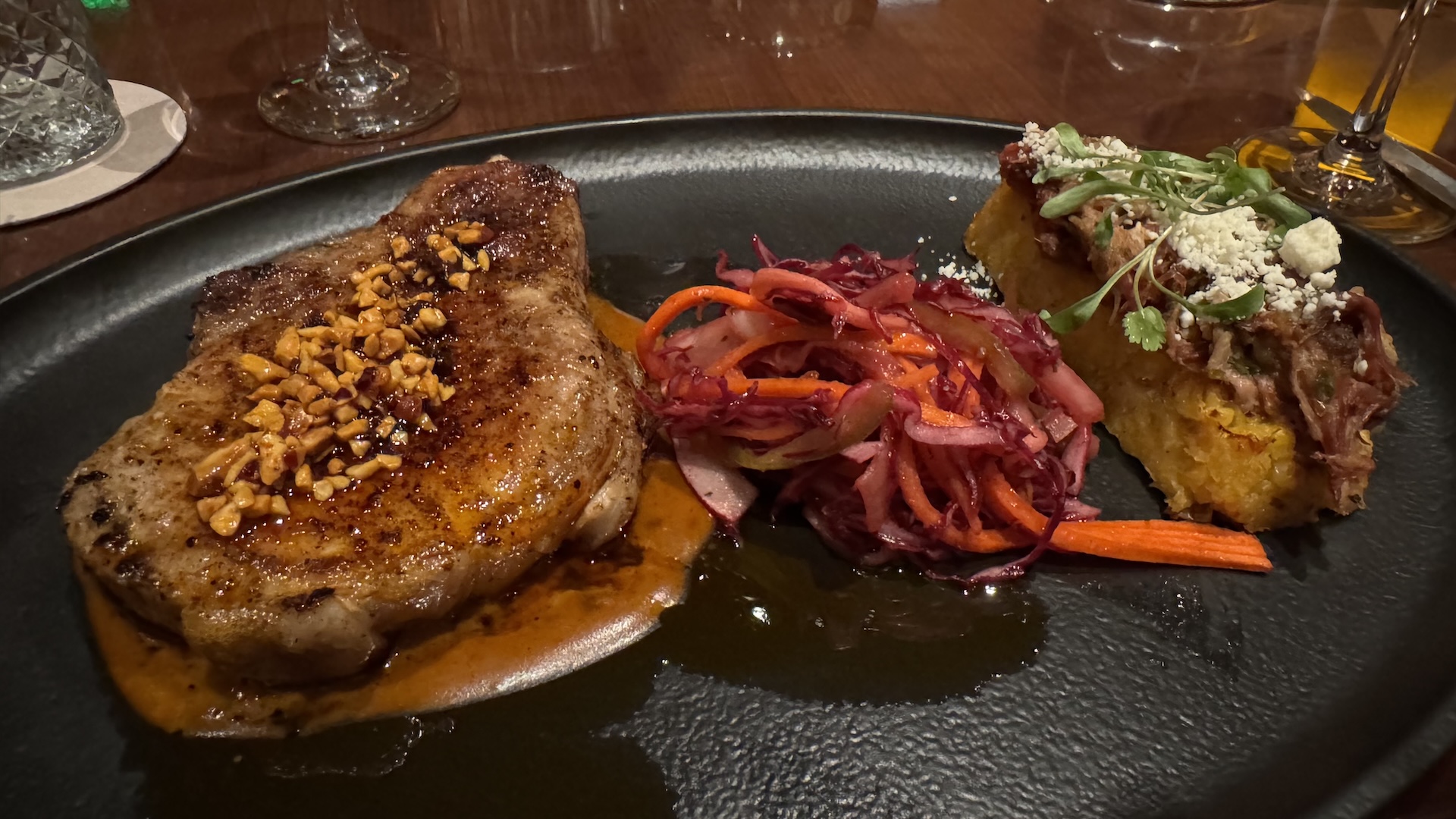 Grilled pork chop with peanuts, slaw, and cornbread on a black plate.