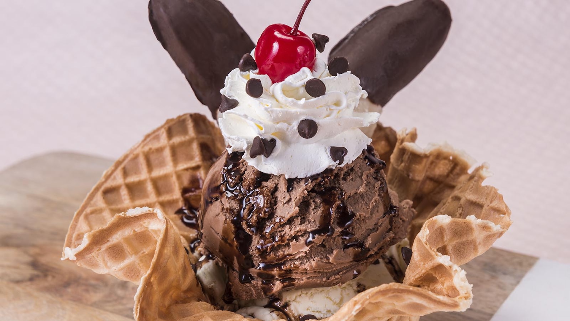 Disney Parks America 250 Foodie Guide The Oswald Sundae: Scoops of vanilla and chocolate ice cream served in a waffle cup topped with whipped cream, hot fudge, chocolate morsels, cherries, and chocolate-dipped frozen bananas