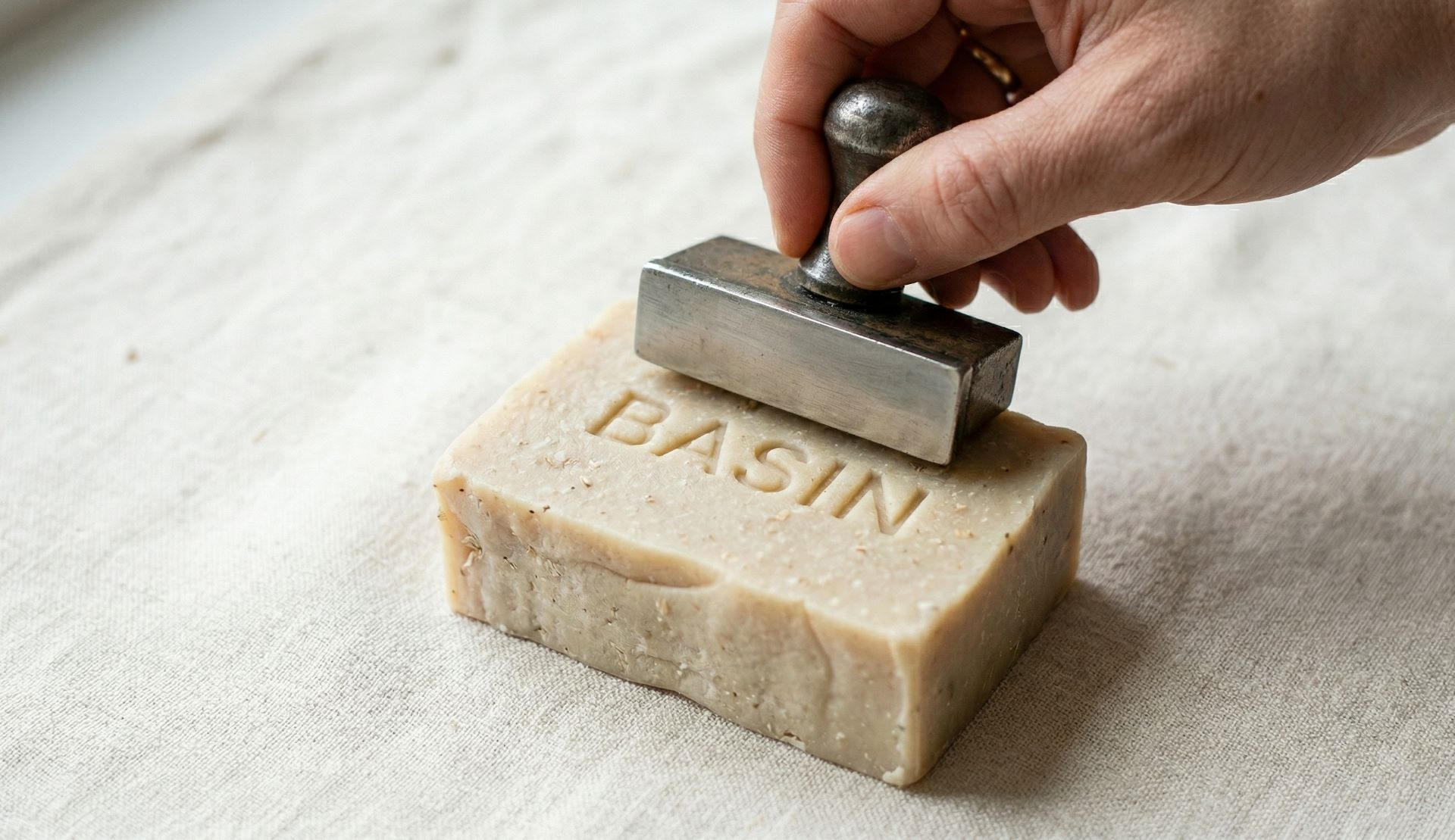 Hand pressing a metal stamp onto a bar of soap labeled "BASIN."