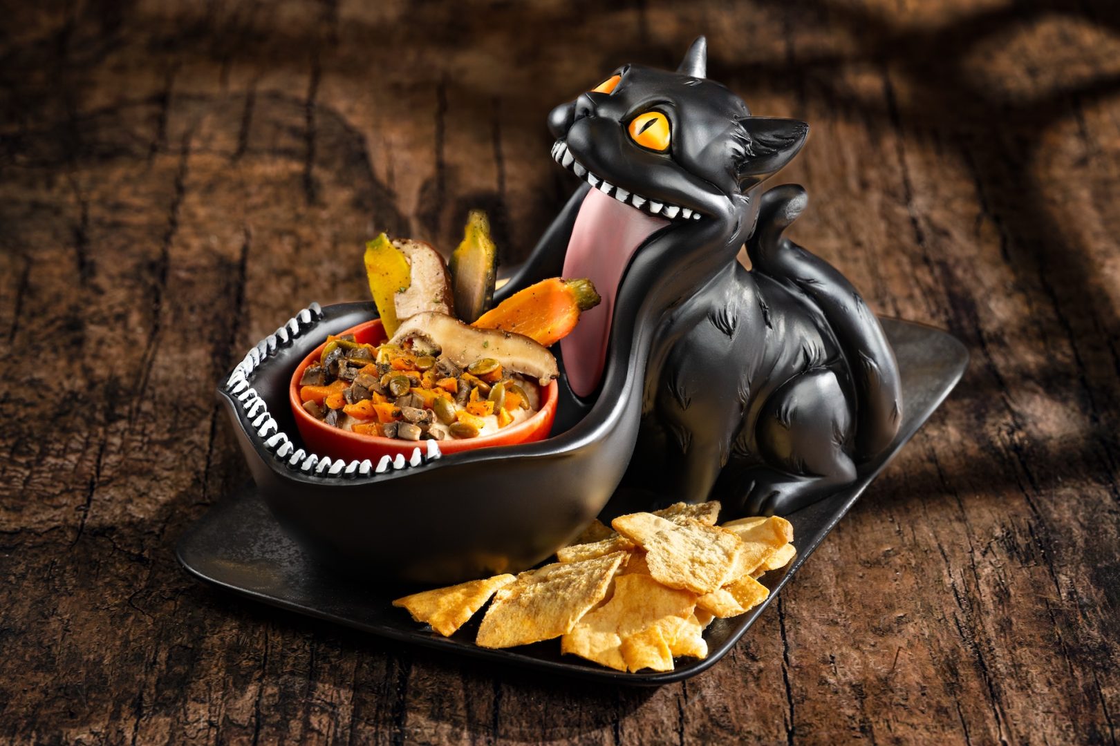 Black cat-shaped dish with dip and chips on a wooden surface.