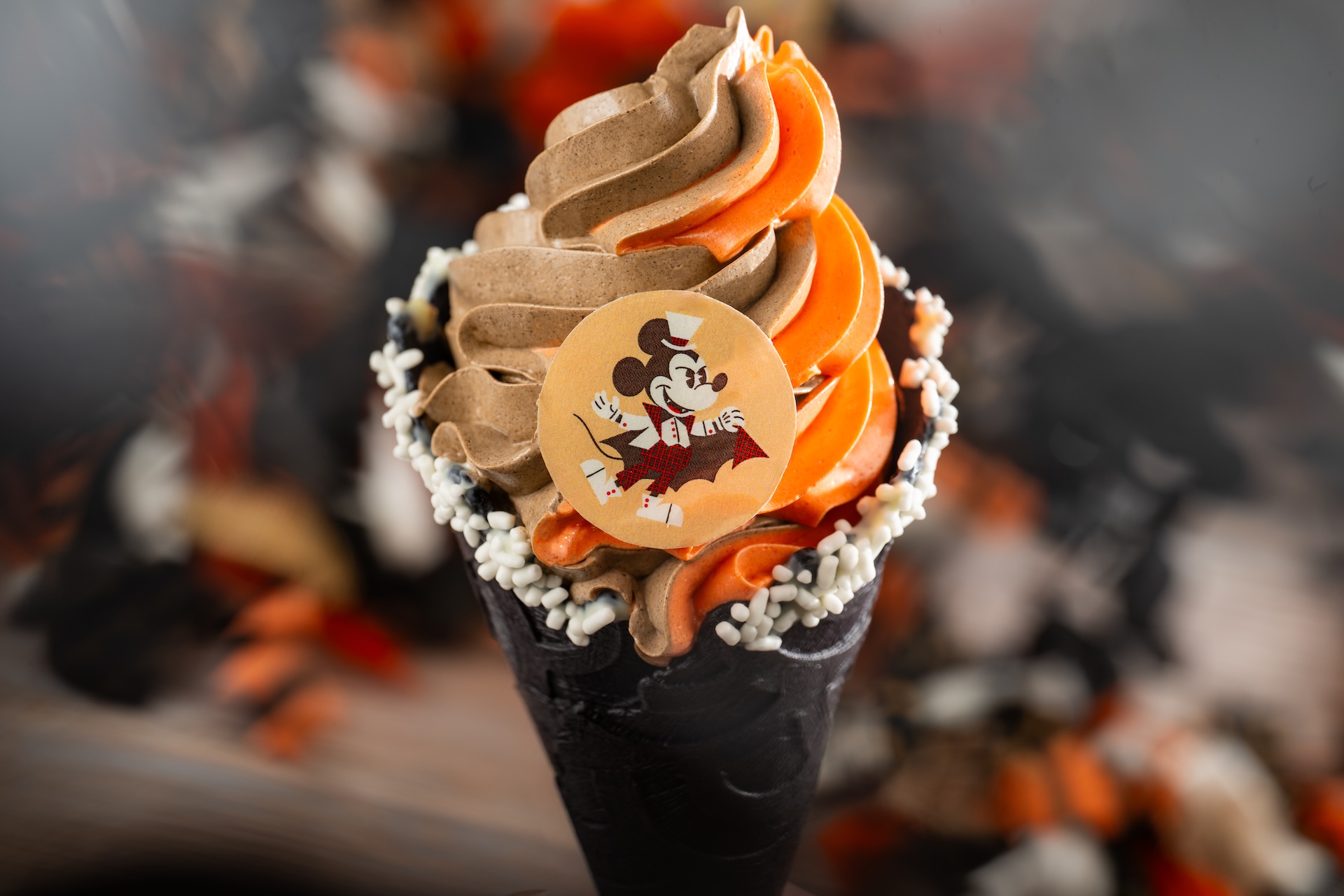 Vampire Mickey Cone: Pumpkin and chocolate soft-serve swirl, white sprinkles, and chocolate Mickey coin in a chocolate cone (New)