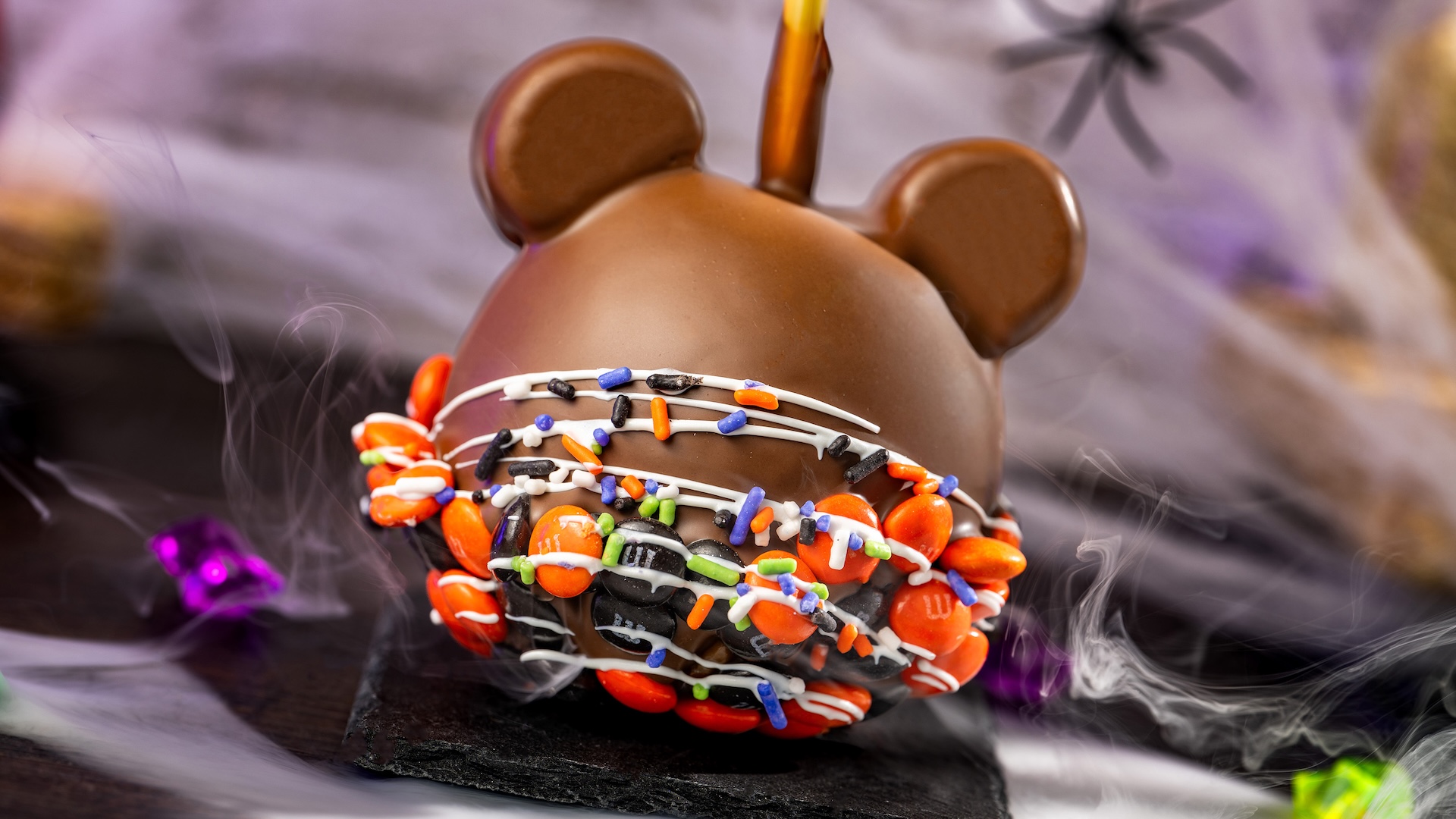 Mickey Halloween Caramel Apple with M&M&rsquo;S Milk Chocolate Candies: Granny Smith apple with marshmallow ears dipped in caramel and milk chocolatey coating with M&M'S milk chocolate candies Halloween blend, Halloween sprinkles, and white chocolatey drizzle 