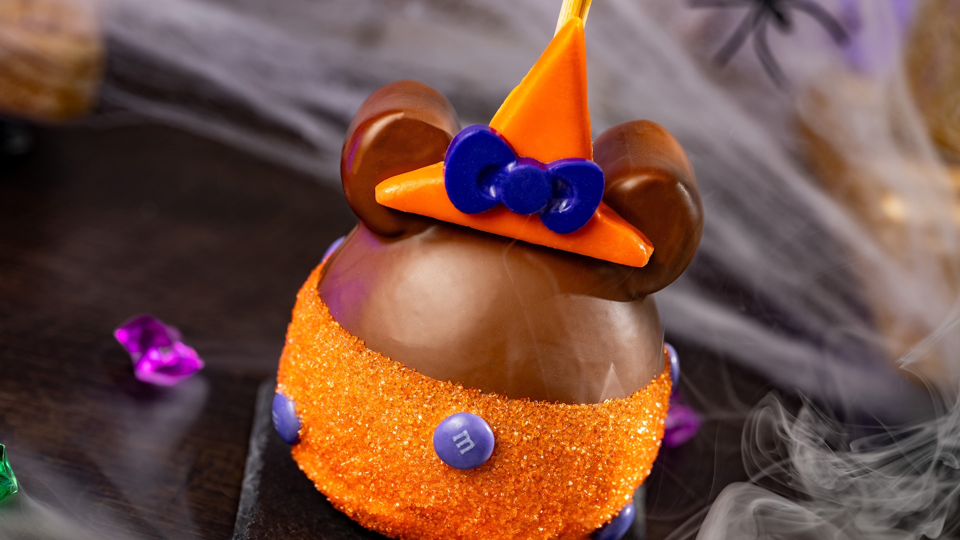 Minnie Witch Caramel Apple with M&M&rsquo;S Milk Chocolate Candies: Granny Smith apple with marshmallow ears dipped in Werther&rsquo;s Original caramel and milk chocolatey coating with M&M'S milk chocolate candies, orange sanding sugar, and a chocolatey witch hat 